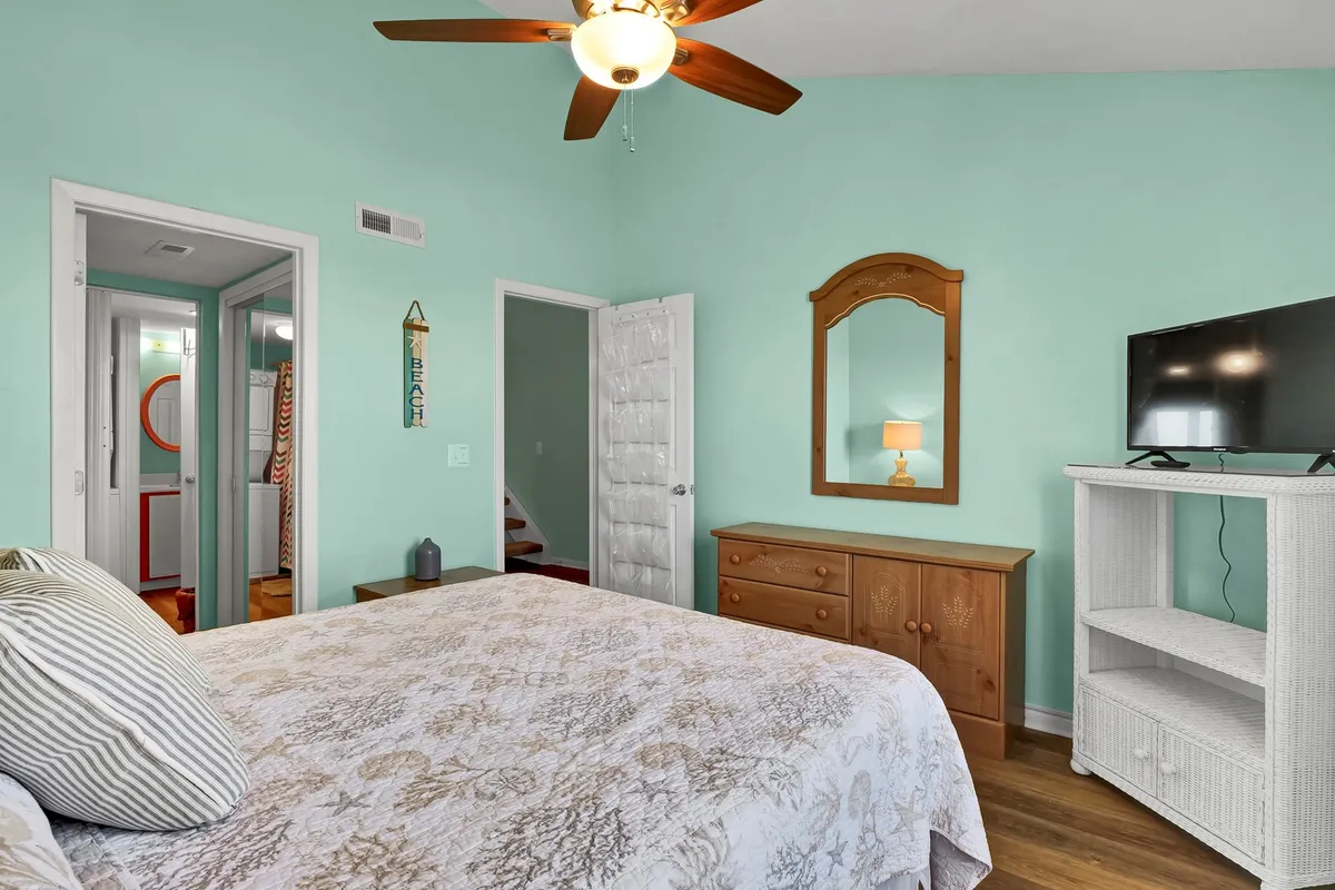 A serene master bedroom is depicted with a comfortable bed, dresser, TV, mirror, and ceiling fan. Natural light filters into the space, highlighting its tranquil and restful atmosphere.
