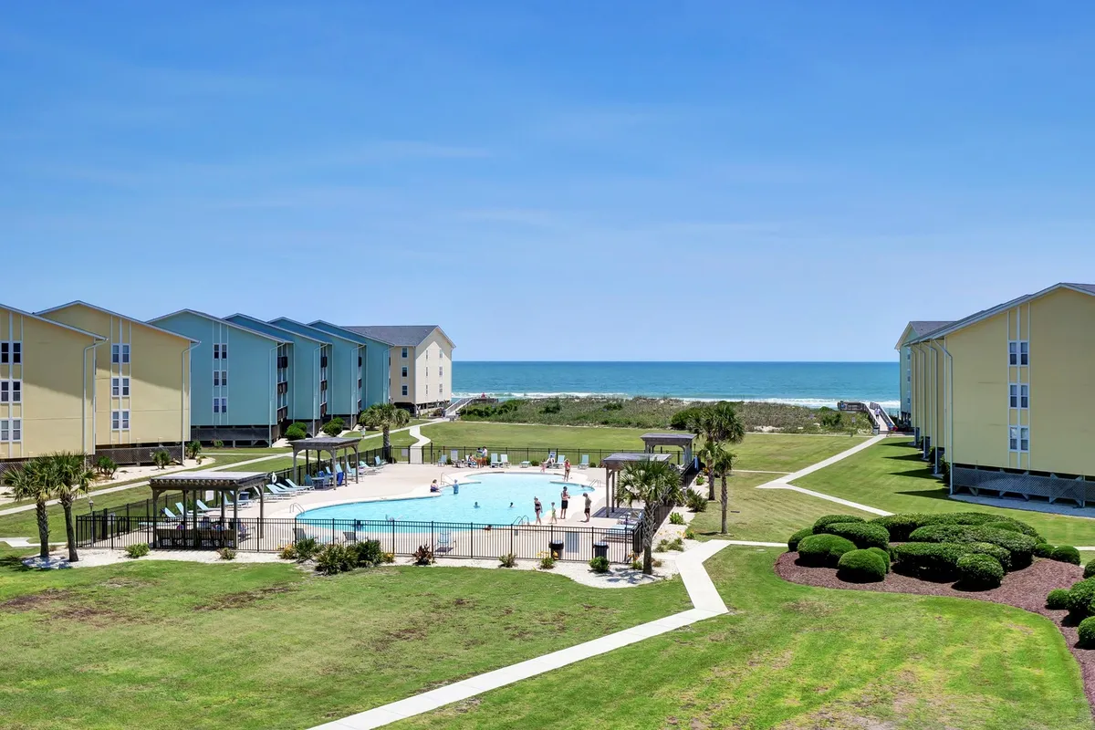 The image showcases the exterior of the Surf Condos community, featuring a sparkling swimming pool with children playing, sunbathers lounging by the water, and a backdrop of sandy beach and ocean waves. The sun is setting over the water, casting a warm, inviting glow, while guests enjoy leisurely walks and barbecue gatherings on the manicured lawn.