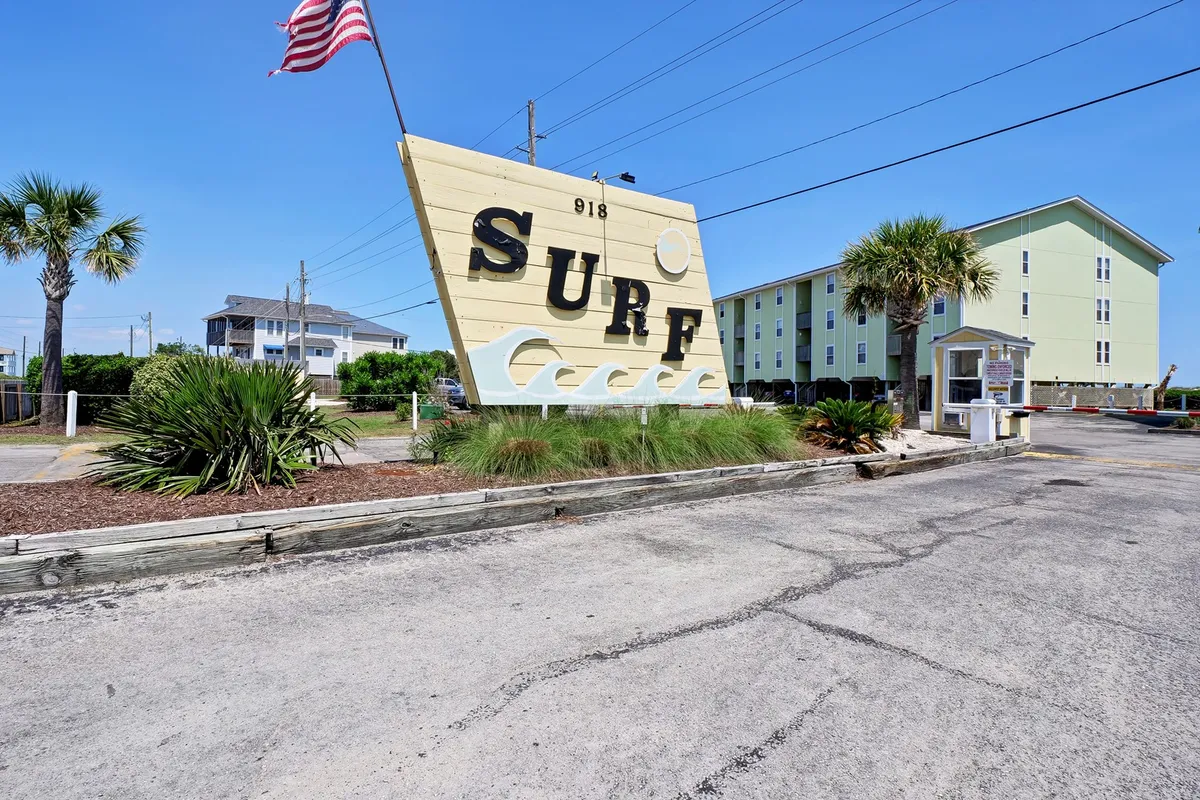 A cheerful exterior scene featuring a large, welcoming 'SURF' sign, surrounded by palm trees and neighborhood buildings. The area exudes a beach-town vibe, hinting at lively exploration and proximity to local attractions.