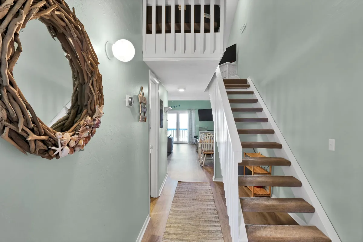 This image captures the inviting entryway, featuring a driftwood wreath on the wall, a staircase, and an open doorway leading further into the home. The scene is light-filled and suggests a warm welcome, guiding guests toward the interior spaces.