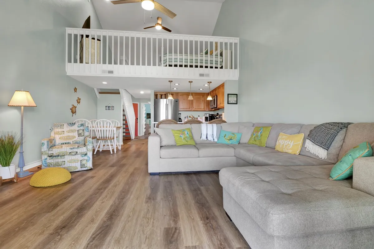 A bright, open-concept living and dining area is shown, with a comfortable sofa adorned with colorful cushions, a dining table, and a visible loft area above featuring a bed. Natural light streams in, and the arrangement captures the playful, flexible family-friendly layout described in the text.