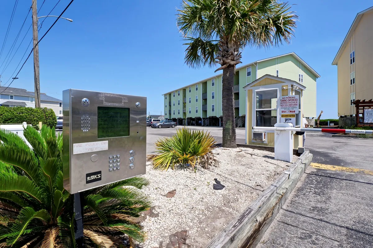 This image depicts the main entrance of the property with lush palm trees, a welcoming entrance booth, and guests socializing outdoors. The intercom system and green building provide context for arrival, while the presence of people conveys an inviting, communal atmosphere.