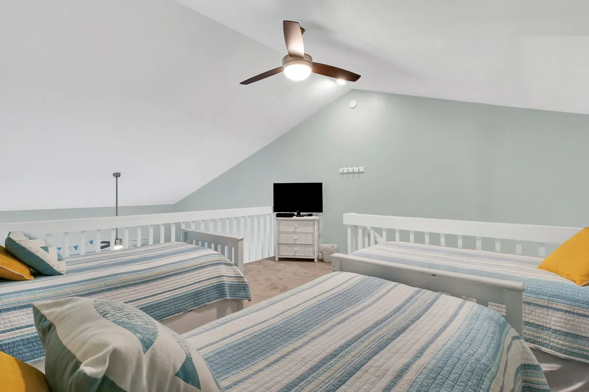A loft area furnished with three twin beds and a television, under a ceiling fan. The setup is relaxed, providing a playful, shared space perfect for kids or friends to unwind together, echoing the loft description in the section.