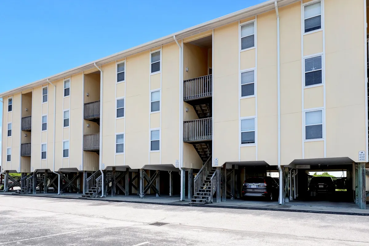 The building structure, visible staircase, and open parking area are shown, with groups of people casually socializing outside. The sunny setting and landscaped grounds invite families and friends to gather and enjoy time together outdoors.
