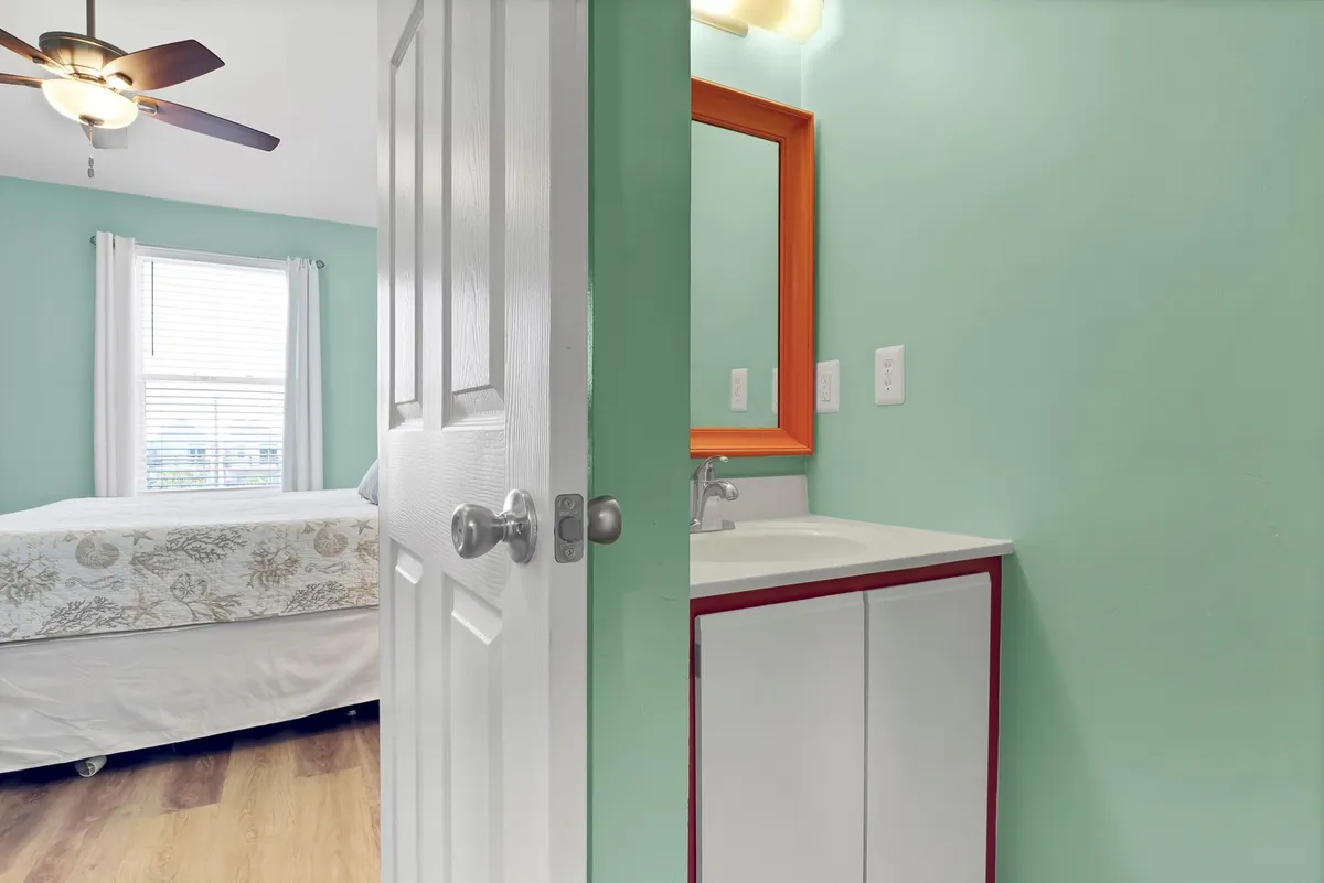 The image reveals a bedroom with a view into the attached bathroom, featuring a colorful orange mirror and a bright, clean sink area. The space feels light-hearted and modern, offering a glimpse of the en-suite convenience.