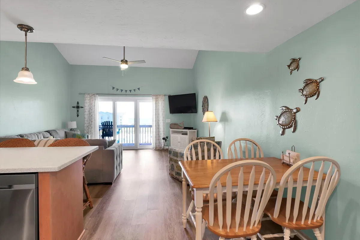 An interior shot from the living room looking out through sliding glass doors onto the deck, showing a dining table, sofa, and cheerful décor. The deck and view beyond are visible, blending the indoor and outdoor living experience.