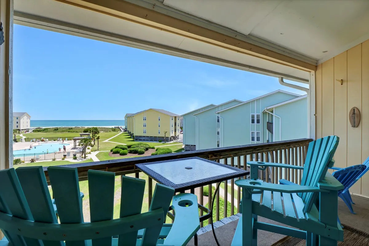 This image shows blue Adirondack chairs positioned on a private balcony or deck, overlooking both the community pool and the vast Atlantic Ocean beyond. The balcony railing frames an inviting view, and the scene conveys a tranquil space perfect for relaxing with a drink or enjoying the fresh sea breeze.