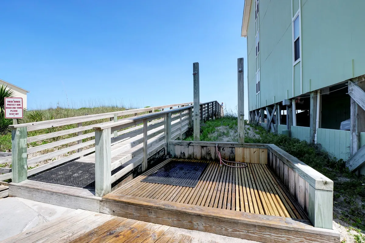 A wooden deck pathway leading to a shower area, ideal for rinsing off after the beach or pool. The setting is coastal, with breezes and a relaxed vibe, and clearly highlights the convenience of on-site shower and foot wash stations.