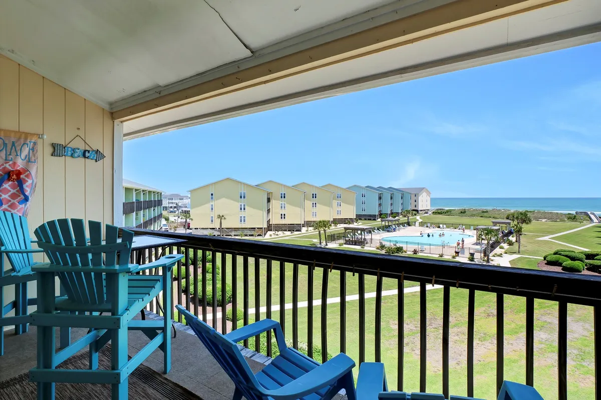 An inviting exterior shot of a balcony with blue Adirondack chairs overlooking the sparkling pool, colorful buildings, and the ocean in the distance. The view captures community and natural beauty in a single frame, emphasizing space for relaxation and gatherings.
