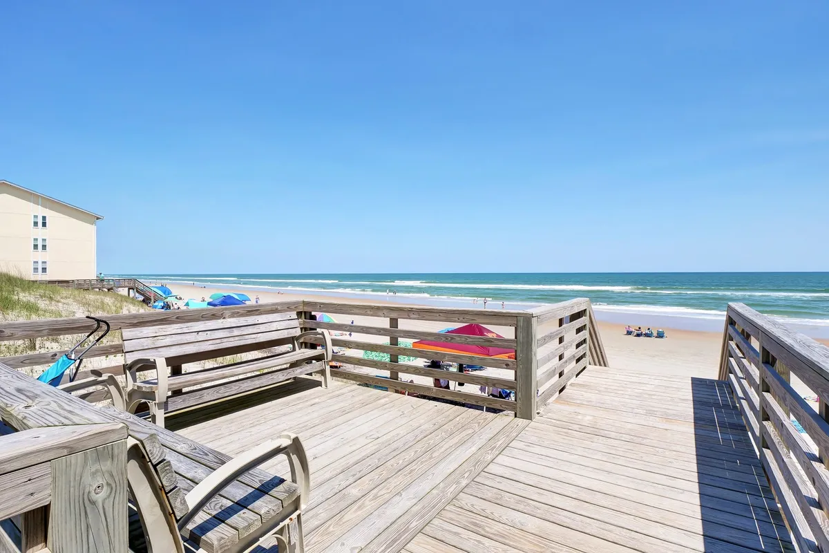 A wide wooden deck overlooks the sparkling Atlantic Ocean, with beach umbrellas providing pops of color. The scene invites relaxation and socializing, and the ocean is just steps away, hinting at easy beach access and the possibility of family gatherings on the deck.